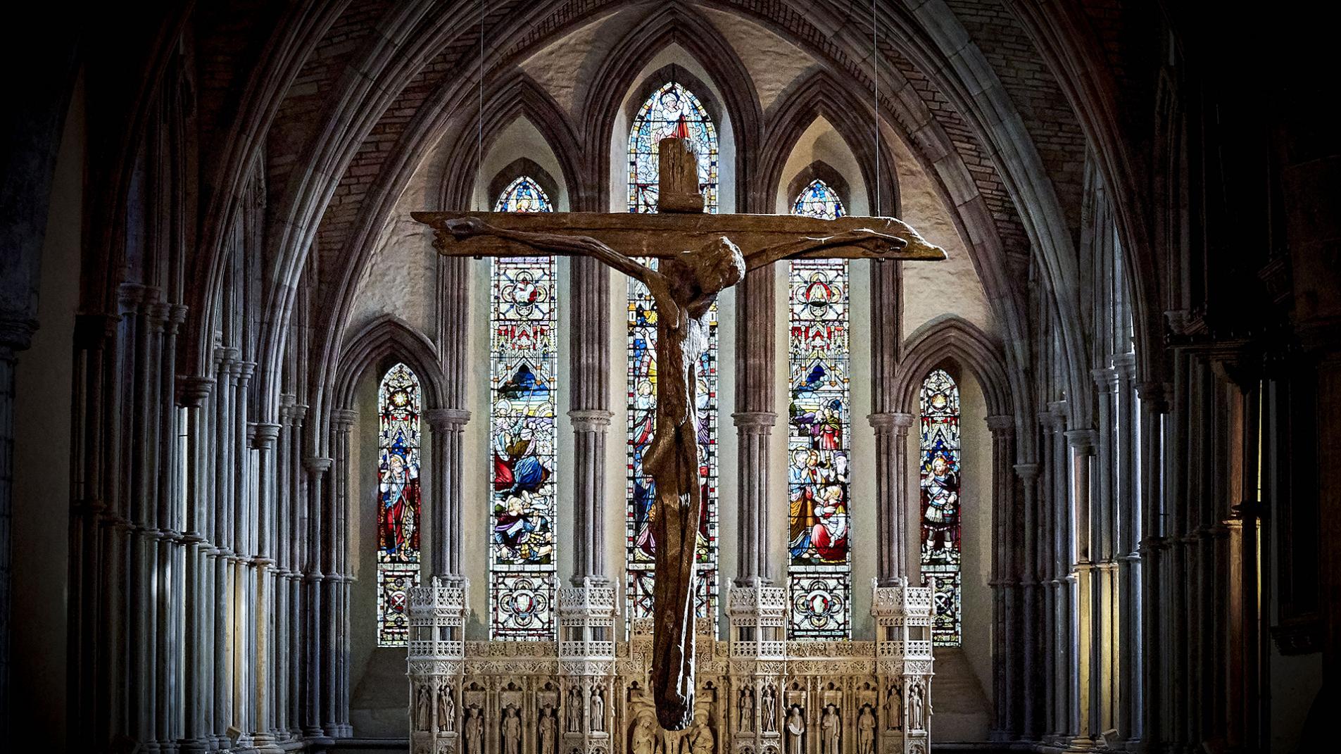 Image of interior of Brecon Cathedral, stain glass window in background with wooden cross in foreground