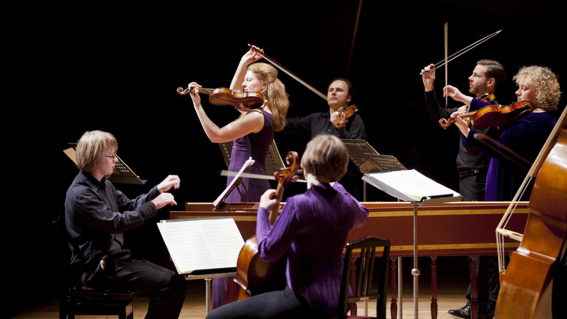 Brecknock Sinfonia, i the foreground is a man playing a harpsichord, a female cellist has her back towards the camera. In the background are four violinists, violins on their shoulders and bow held in the air